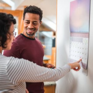 Good looking multiracial pregnant couple pointing at a calendar and counting the days to receive their first child. They are talking and bonding.